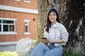 An attractive young Asian female student is sitting and relaxing under the tree in a campus park Royalty Free Stock Photo