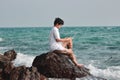 Attractive young Asian man with laptop sitting on the rock of turquoise sea shore. Happy vacations time concept.