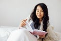 An attractive young Asian woman is lying in bed on pillows and reading a book. A enchanting female Korean is resting on the sofa