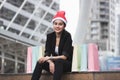 Attractive young Asian woman with santa hat and colorful shopping bag sitting outdoors after shopping for christmas gifts Royalty Free Stock Photo
