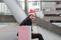 Attractive young Asian woman with santa hat and colorful shopping bag sitting outdoors after shopping for christmas gifts Royalty Free Stock Photo
