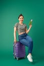 Attractive young Asian woman sitting on suitcase and holding passport, ticket flight over green background
