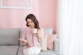 Attractive young Asian woman using phone and drinking coffee when sitting on sofa at home Royalty Free Stock Photo