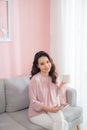 Attractive young Asian woman using phone and drinking coffee when sitting on sofa at home Royalty Free Stock Photo