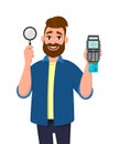 Attractive young bearded man showing/holding magnifying glass and credit/debit card swiping machine or POS terminal. Search, find. Royalty Free Stock Photo