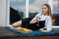 Attractive young blond woman in black dress holding wineglass, drink champagne and eat fruits, lying on blanket. Relax Royalty Free Stock Photo