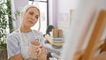 Attractive young blonde artist thoughtfully looking at drawing while sipping coffee in art studio, array of paintbrushes at her Royalty Free Stock Photo