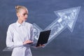 Attractive young blonde european businesswoman with laptop and upward business arrow sketch drawing on chalkboard wall background Royalty Free Stock Photo