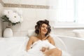 Attractive young brunette woman in hair curlers enjoying her bath with bubbles in bright bathroom