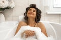 Attractive young brunette woman in hair curlers enjoying her bath with foam in bright bathroom