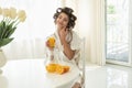 Attractive young brunette woman in hair curlers enjoying orange fresh squeezed juice sitting in bright kitchen having Royalty Free Stock Photo