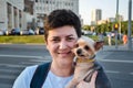 Attractive young brunette woman with short hair holds in her arms small cute Yorkshire terrier dog on background of city street.