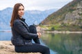 Attractive young brunette woman sits on stone pier and gazes thoughtfully out to sea, beautiful mountain range on Royalty Free Stock Photo