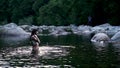 An attractive young brunette woman swimming in a pristine forest river with a creepy voyeur photographing her from afar.