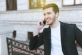 Attractive young business man using mobile phone on building outdoors. Cheerful handsome caucasian man in black suit in the city.