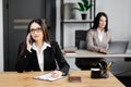 Attractive young business woman wearing jacket talking on mobile phone while sitting on a desk and using notebook near her Royalty Free Stock Photo