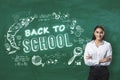 Attractive young businesswoman with folded arms standing on creative back to school sketch on blackboard background. Education,