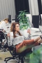 Attractive young businesswoman holding cup and using smartphone while sitting