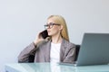 Attractive young businesswoman wearing jacket talking on mobile phone while sitting on a desk and using laptop computer in office Royalty Free Stock Photo