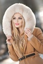 Attractive young Caucasian adult with light brown fur coat hood. Beautiful blonde girl with gorgeous eyes wearing fur, outdoors