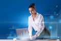 Attractive young caucasian businesswoman leaning on desktop with laptop and abstract glowing business chart hologram on blurry