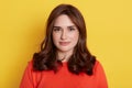 Attractive young Caucasian dark haired female posing against yellow background, dressed in casual orange sweater with calm face