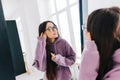 Attractive young caucasian woman in eyeglasses touching face and looking at mirror in living room at home in sunny day