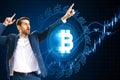 Attractive young european businessman celebrating success with creative glowing bitcoin on dark blue background with candlestick.