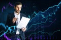 Attractive young european businessman with document using creative glowing candlestick forex chart hologram on blurry dark blue Royalty Free Stock Photo