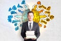 Attractive young european businessman holding books with creative drawing of different brain hemispheres on concrete wall