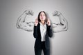 Attractive young european businesswoman with crossed fingers standing on concrete wall background with drawn muscle hands. Royalty Free Stock Photo