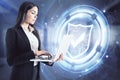 Attractive young european businesswoman with laptop and glowing shield hologram standing in blurry office interior. Innovation, Royalty Free Stock Photo