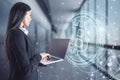 Attractive young european businesswoman using laptop in blurry office interior with glowing bitcoin hologram. Cryptocurrency and Royalty Free Stock Photo