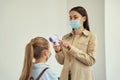 Attractive young female teacher wearing protective mask screening schoolgirl using digital thermometer while kid coming Royalty Free Stock Photo