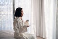 Attractive young girl in bathrobe holding a cup, looking at window and smiling while sitting on a bed near the big Royalty Free Stock Photo