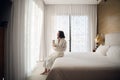 Attractive young girl in bathrobe holding a cup, looking at window and smiling while sitting on a bed near the big Royalty Free Stock Photo