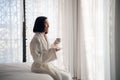 Attractive young girl in bathrobe holding a cup, looking at window and smiling while sitting on a bed near the big Royalty Free Stock Photo