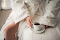 Attractive young girl in bathrobe holding a cup, looking at window and smiling while sitting on a bed near the big Royalty Free Stock Photo