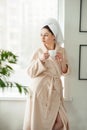 Attractive young girl in bathrobe and with a towel on her head is holding a cup, sitting on a window sill in the bathroom Royalty Free Stock Photo