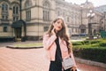 Attractive young girl with long curly hair and red lips posing in city. She wears black dress, pink jacket, clutch bag Royalty Free Stock Photo