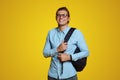 Attractive young guy in blue shirt and eyeglasses holding backpack and smiling at camera on yellow backdrop Royalty Free Stock Photo