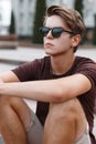 Attractive young hipster man in light shorts in stylish sunglasses with a stylish hairstyle in a trendy T-shirt sits Royalty Free Stock Photo