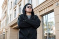 Attractive young hipster man in a stylish black hooded sweatshirt in a fashionable cap in sunglasses poses near a vintage building Royalty Free Stock Photo