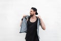 Attractive young hipster man in stylish denim vest in black t-shirt in trendy cap poses near white vintage wall outdoors. Nice guy Royalty Free Stock Photo