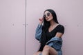 Attractive young hipster woman with black hair in stylish sunglasses in a fashionable denim jacket in a trendy top posing Royalty Free Stock Photo