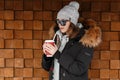 Attractive young hipster woman in knitted vintage hat in sunglasses in a black jacket with a fur hood in a stylish sweatshirt Royalty Free Stock Photo