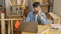 Attractive young hispanic carpenter engrossed in work, talking on smartphone while accessing computer in his carpentry workshop