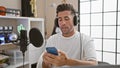 Attractive young hispanic man, a dynamic radio reporter, professionally speaking passionately on air, reading news from smartphone Royalty Free Stock Photo