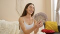 Attractive young hispanic woman smiling, flaunting her wealth as she holds dollar banknotes, sitting in the comfort of her home\'s