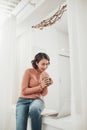 Attractive young lady with cup sitting on window sill at home Royalty Free Stock Photo